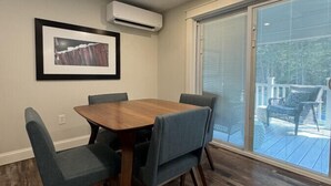 A dining area with a wooden table and blue upholstered chairs, an air conditioning unit on the wall, and a large sliding glass door leading to an outdoor deck.