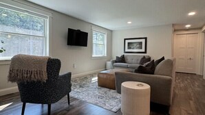 A spacious living room with hardwood flooring, a grey sofa, a patterned armchair, and a small ottoman.