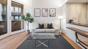 Un salon moderne avec une table basse en verre, un canapé avec des coussins, une plante en pot et un coin cuisine équipé d’un évier et d’un plan de travail.