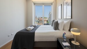 Ein Hotelzimmer mit Bett, Nachttisch mit Lampe, einer Wasserflasche und Blick auf die Stadt durch große Fenster.