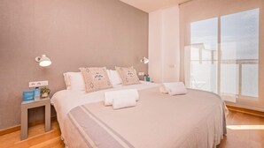 Une chambre à coucher avec un grand lit, deux lampes de chevet, une table de chevet ornée d’une plante et une fenêtre avec des rideaux légers.
