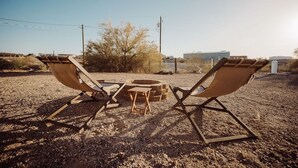 Two lounge chairs facing a fire pit in a desert setting with sparse vegetation and a building in the background. - Secluded and Secure, Pet-friendly Desert Getaway. Your vacation awaits! (Lake Havasu City)