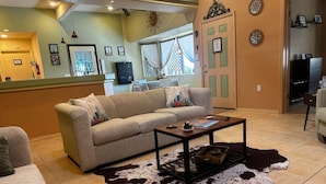 A living room with a sofa, a coffee table, and a cowhide rug. There is a door with a sign, a window with curtains, and a wall with framed pictures. - Secluded and Secure, Pet-friendly Desert Getaway. Your vacation awaits! (Lake Havasu City)