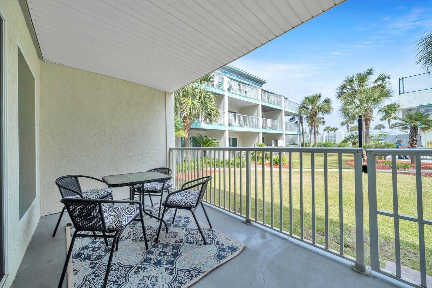 A balcony with a table and chairs, a patterned rug, and a view of a multi-story building with balconies and palm trees.