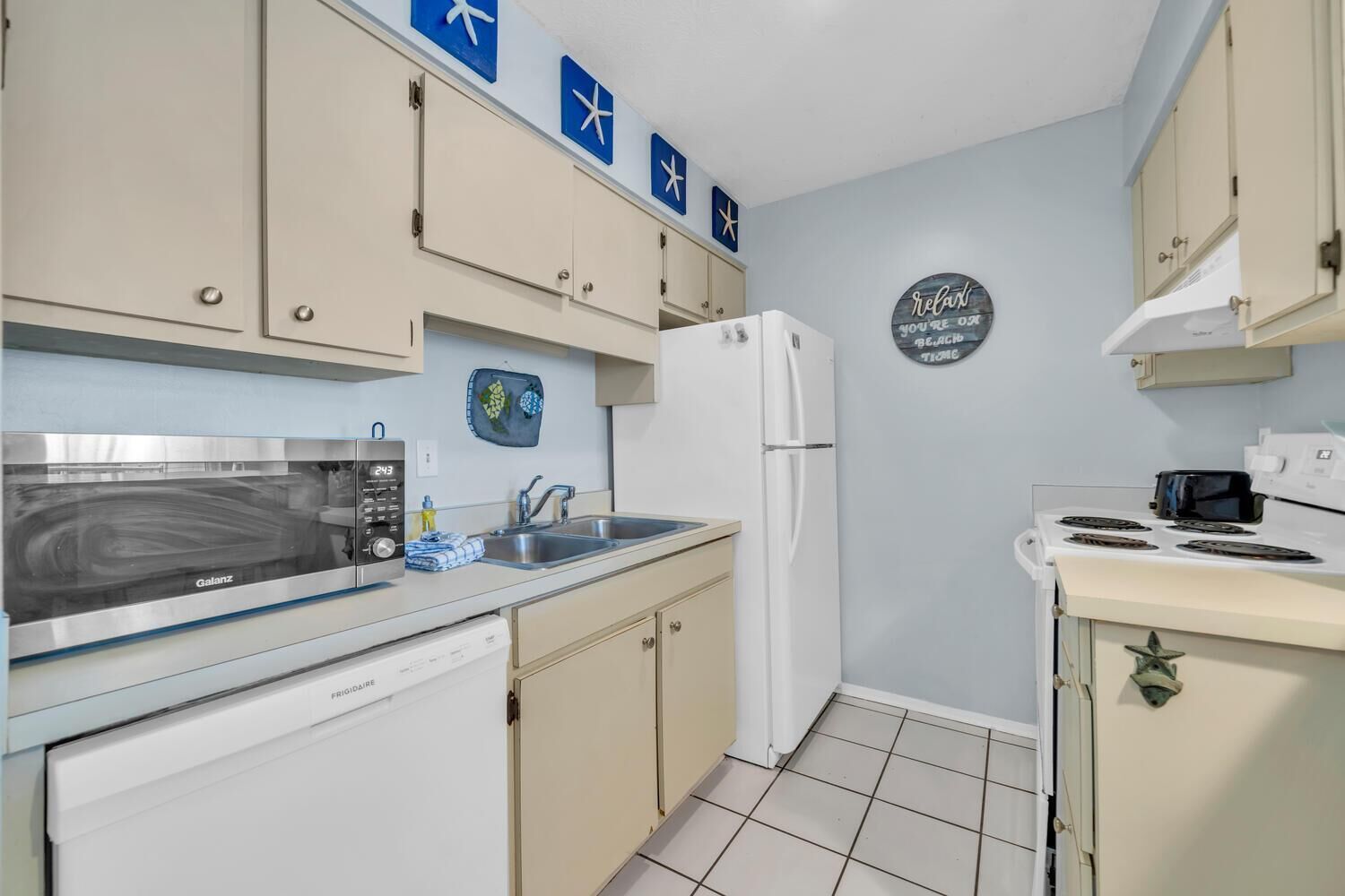 A compact kitchen with a Galanz microwave, a white refrigerator, and a stove. There are cabinets, a sink, and a decorative sign on the wall.