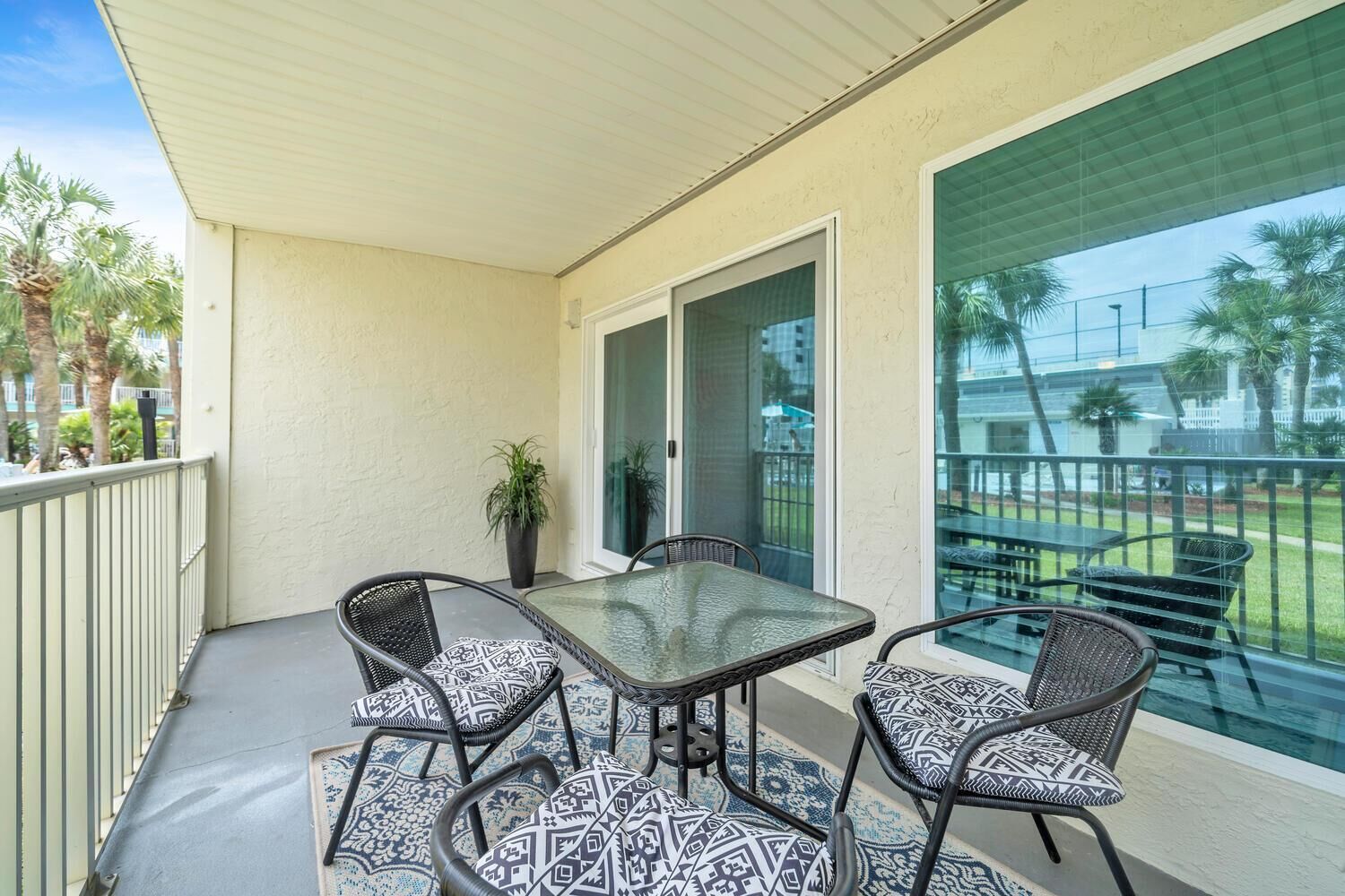 A covered balcony with a glass table and four chairs, a potted plant, and a view of a pool and palm trees.