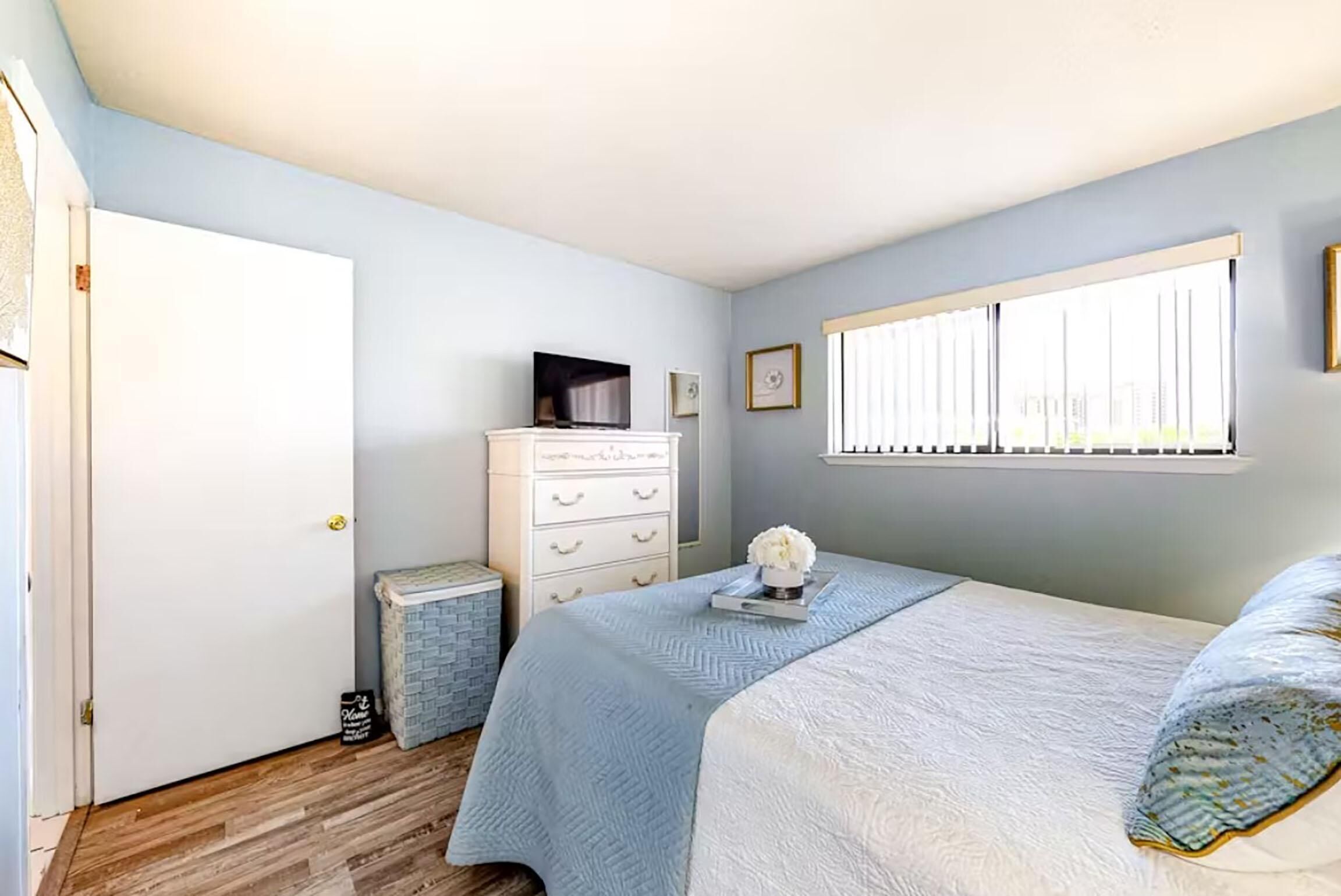 Een slaapkamer met een bed, een dressoir, een televisie en een raam met jaloezieën.