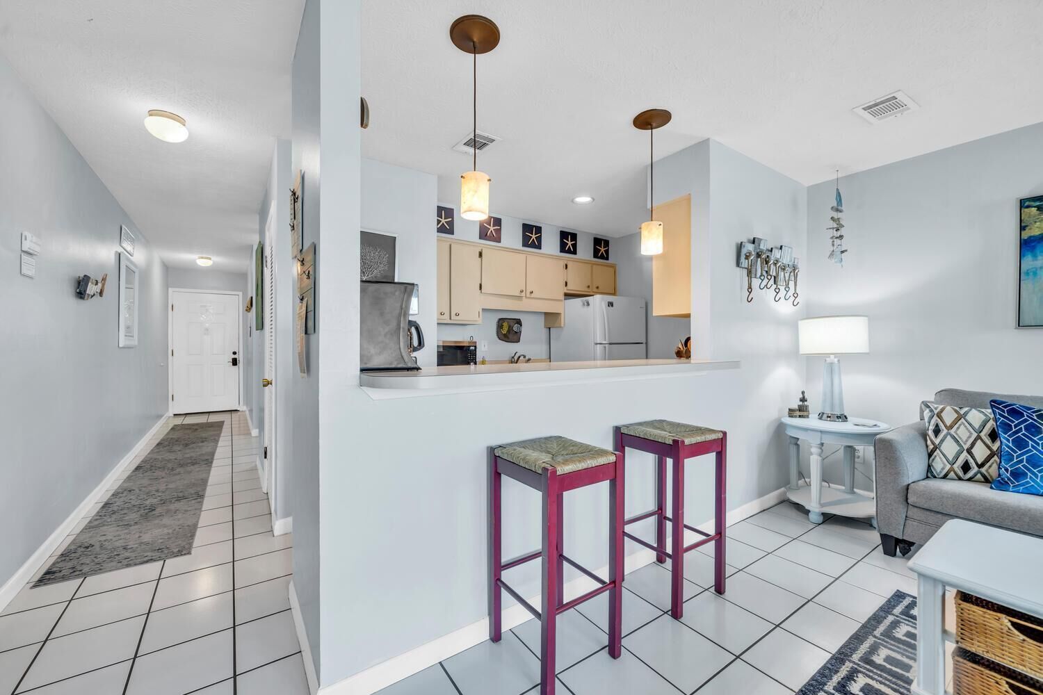 Une cuisine moderne avec un îlot central, deux tabourets de bar, une petite table avec une lampe et un canapé avec des coussins.