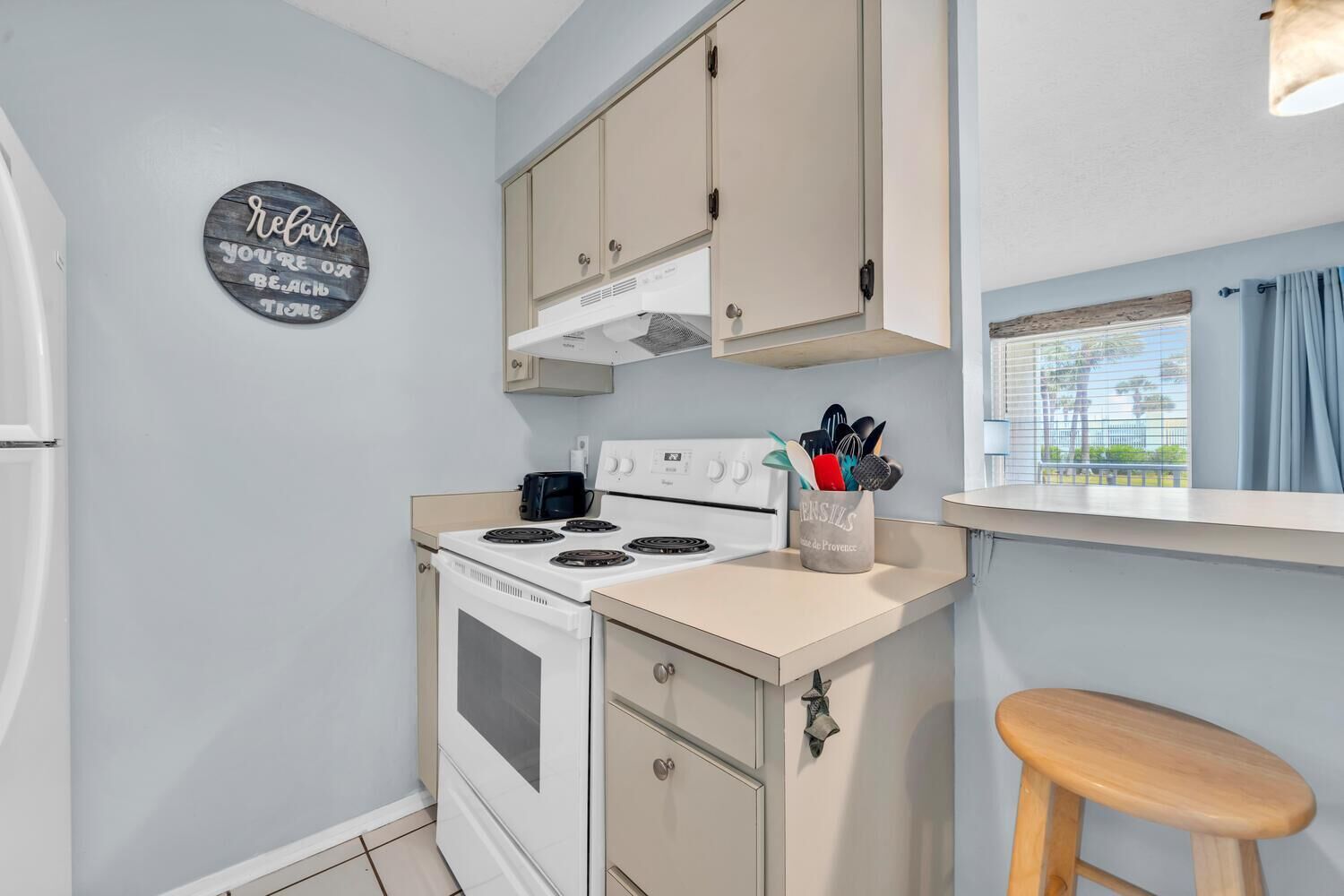 A compact kitchen with a white stove, beige cabinets, and a wooden stool. A sign on the wall reads "Relax, you're on vacation."