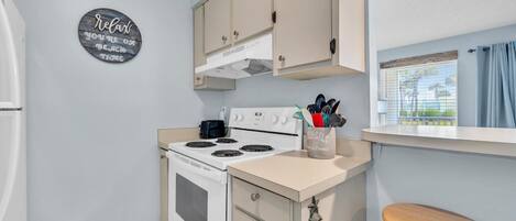A compact kitchen with a white stove, beige cabinets, and a wooden stool. A sign on the wall reads "Relax, you're on vacation."