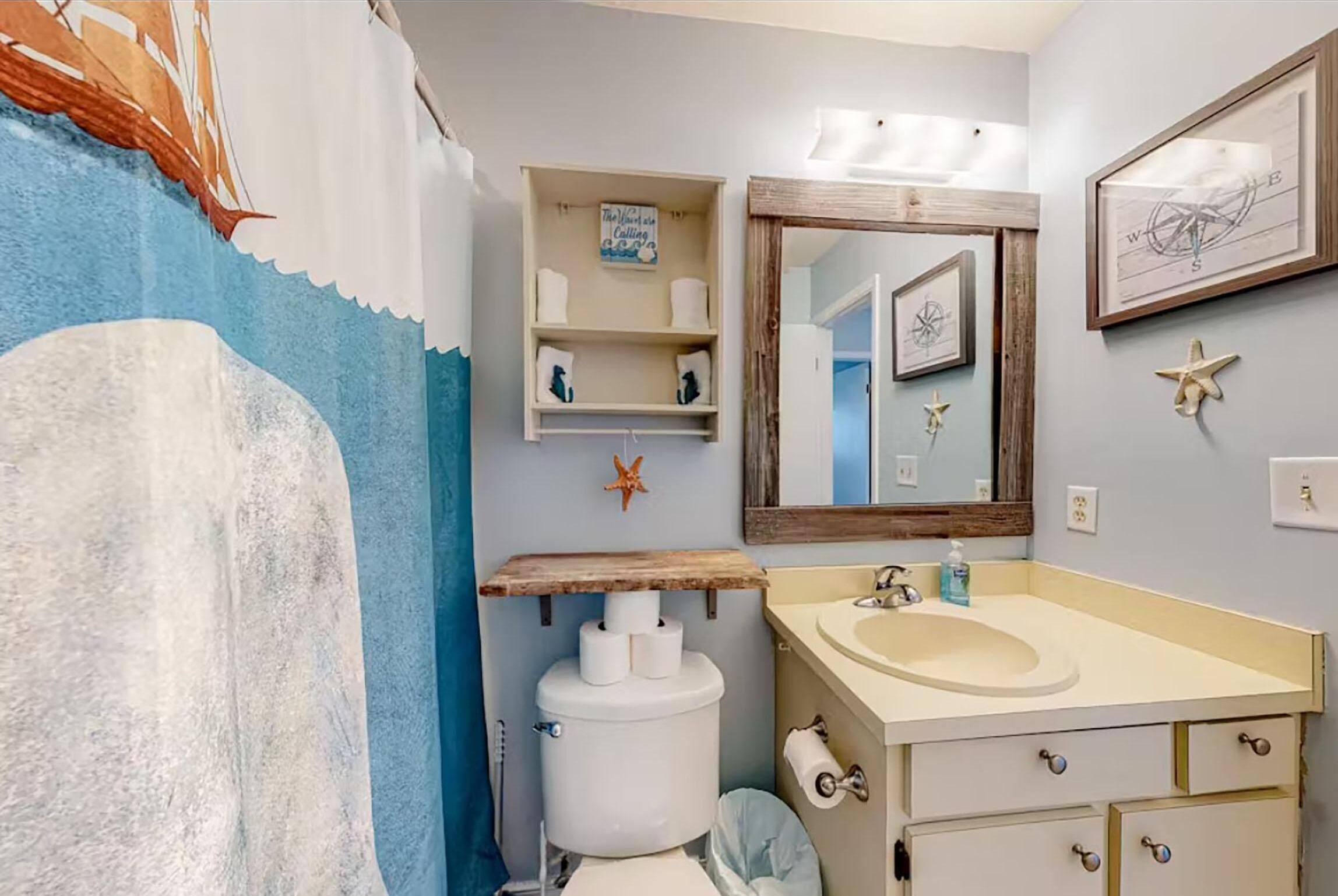 Un baño con temática náutica, que presenta un gran mural de barco, un estante de madera con artículos de aseo, un lavamanos redondo y un espejo con diseño de brújula.