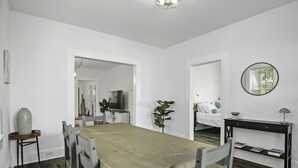 A modern, well-lit room with a wooden dining table, chairs, a small side table, a potted plant, a bed with pillows, a bedside table with a lamp, and a wall-mounted mirror. - Park Circle's Blue Ranch                      -Charleston- (North Charleston)