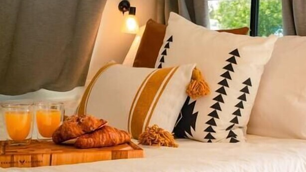 A bed with pillows, a wooden tray with croissants and orange juice, and a window with curtains. - RV Glamping w/ Cowboy Tub, Lawn Games & Firepit (Hastings County)