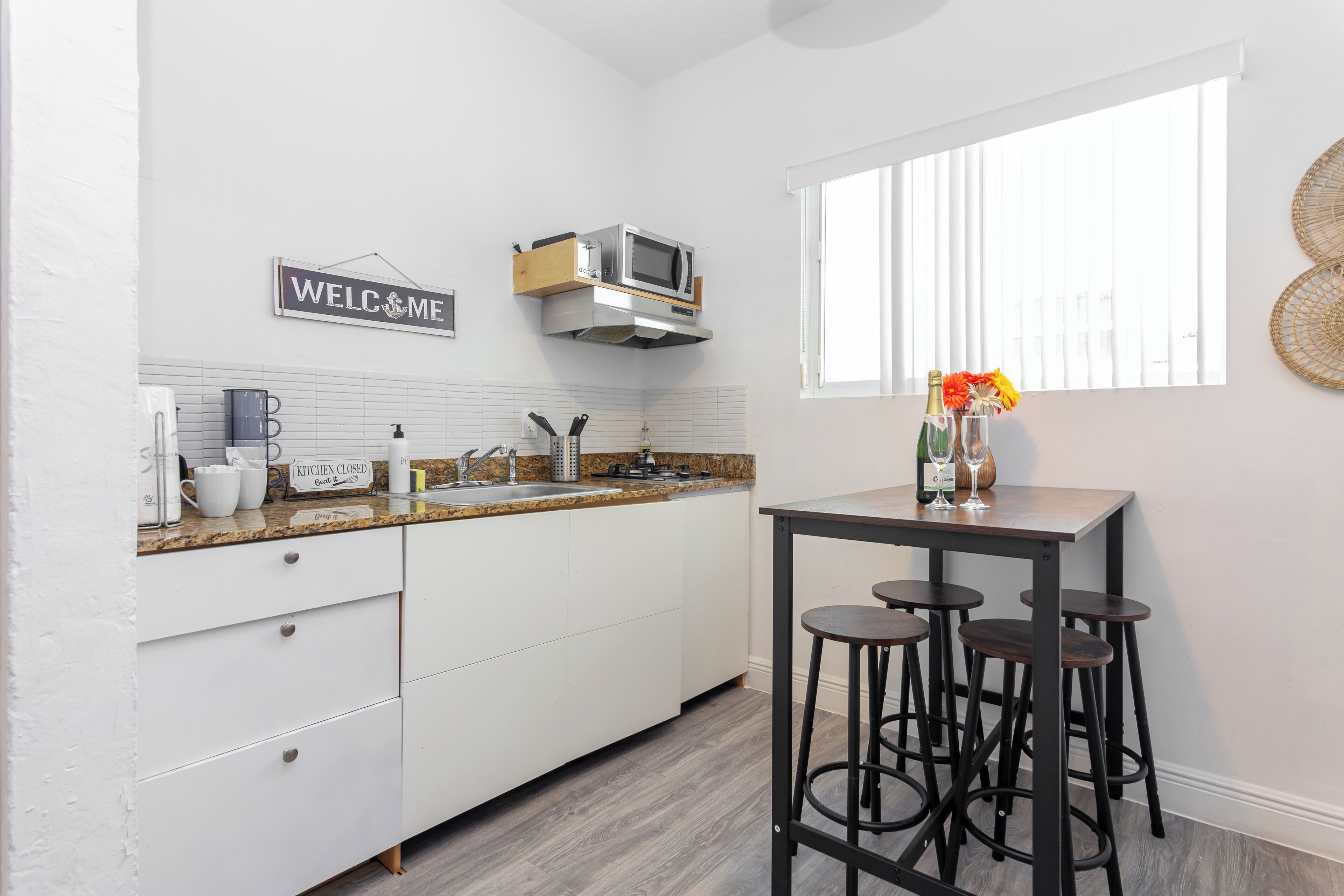 Een moderne keuken met witte kastjes, een granieten aanrecht, een magnetron en een bord met de tekst 'Welcome'. Er is een raam met jaloezieën, een barkruk en een vaas met bloemen op het aanrecht.