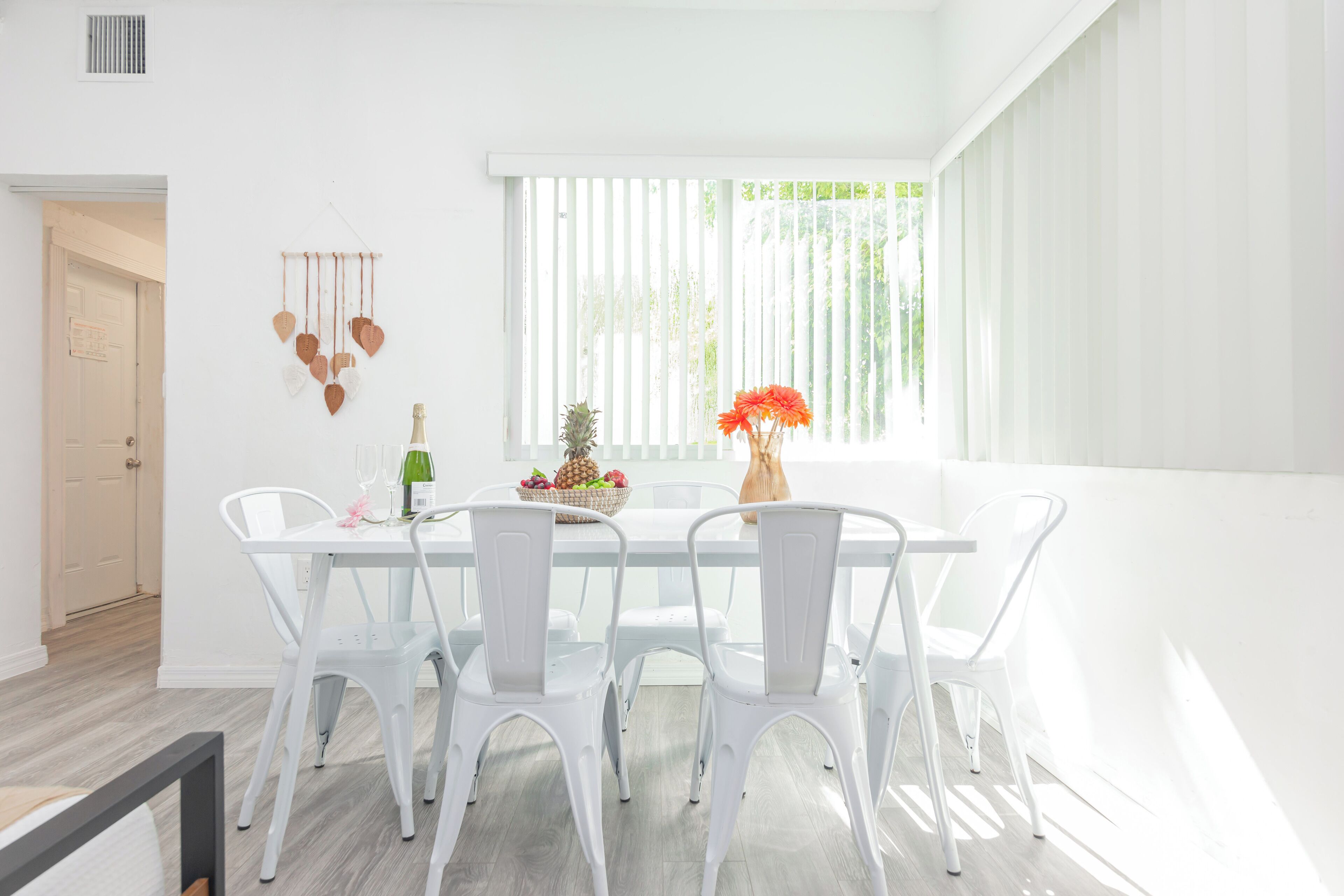 Un comedor luminoso con una mesa y sillas blancas, un adorno con forma de corazón en la pared y una ventana con persianas.