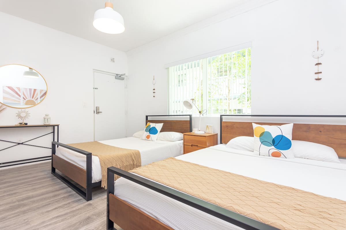 A hotel room with two beds, a desk, a mirror, and a window with blinds.