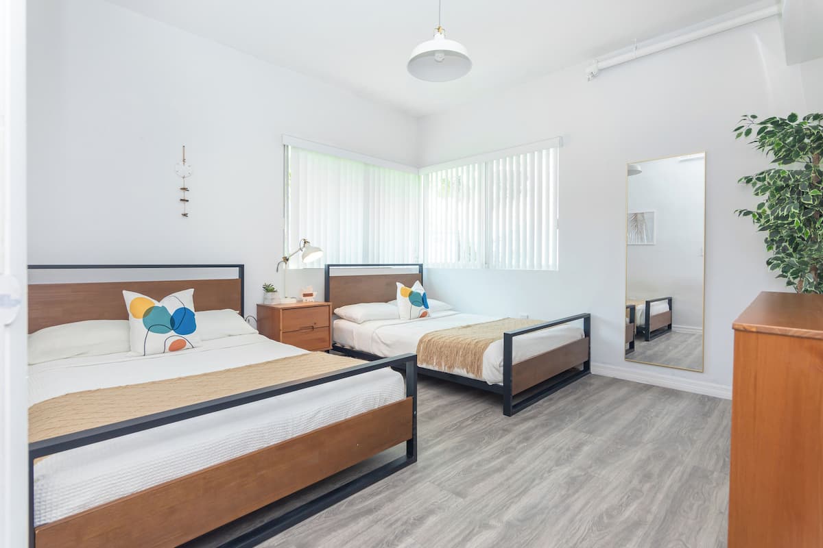 A modern bedroom with two beds, a bedside table with a lamp, a large mirror, and a potted plant.