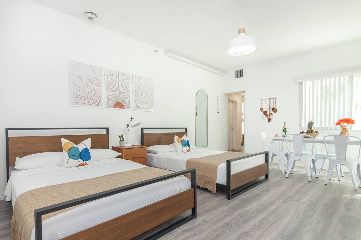 A hotel room with two beds, a dining area, and a large window with blinds.