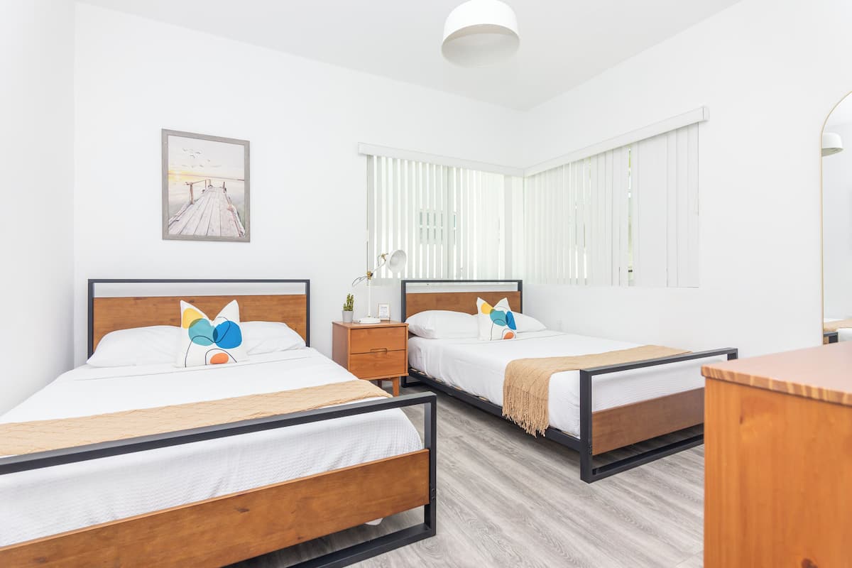 A hotel room with two beds, a wooden floor, a painting on the wall, and a window with blinds.