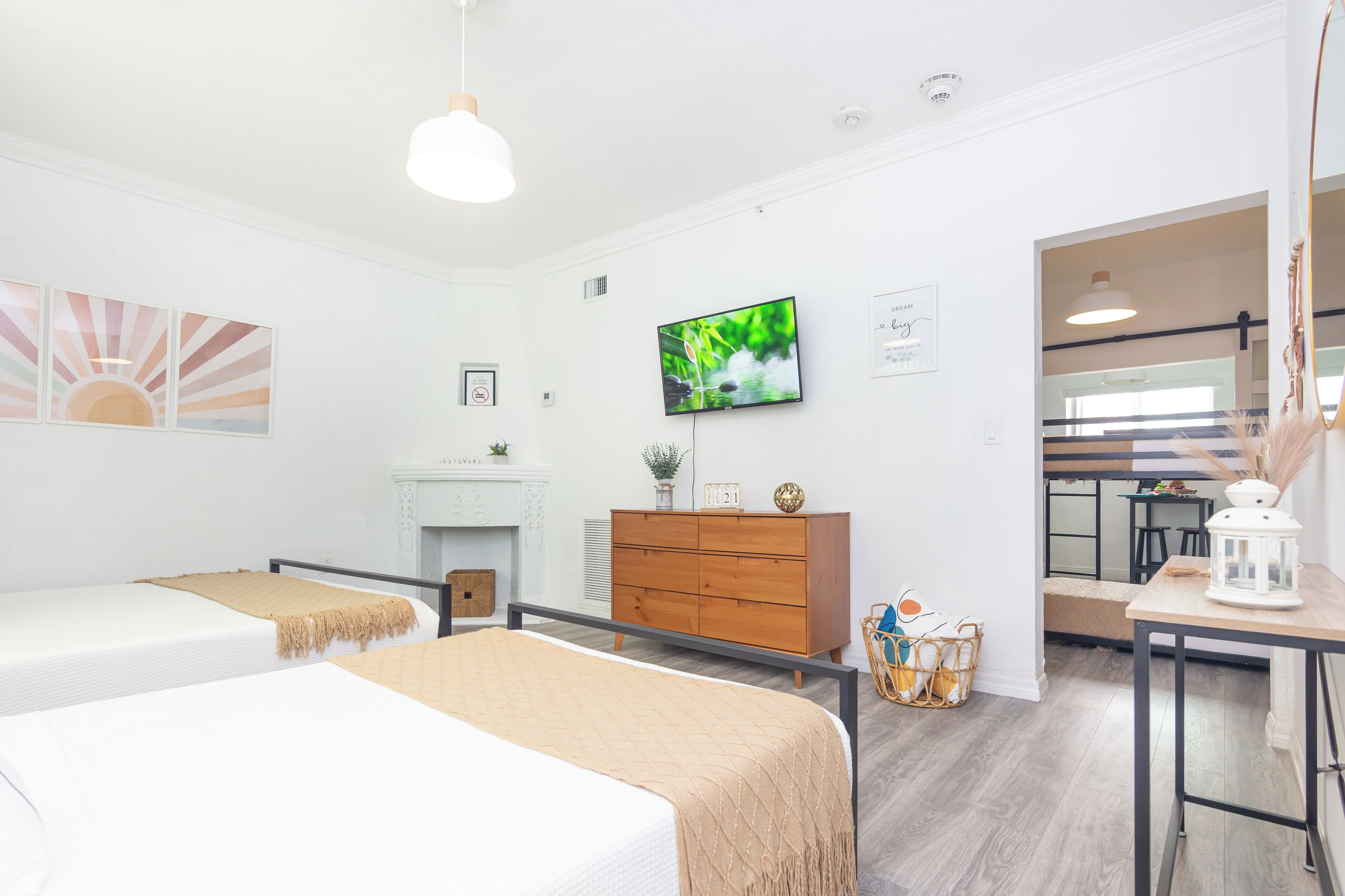 Ein Hotelzimmer mit zwei Betten, einem Fernseher, einer Holzkommode, einem kleinen Tisch und Blick zur Küche.