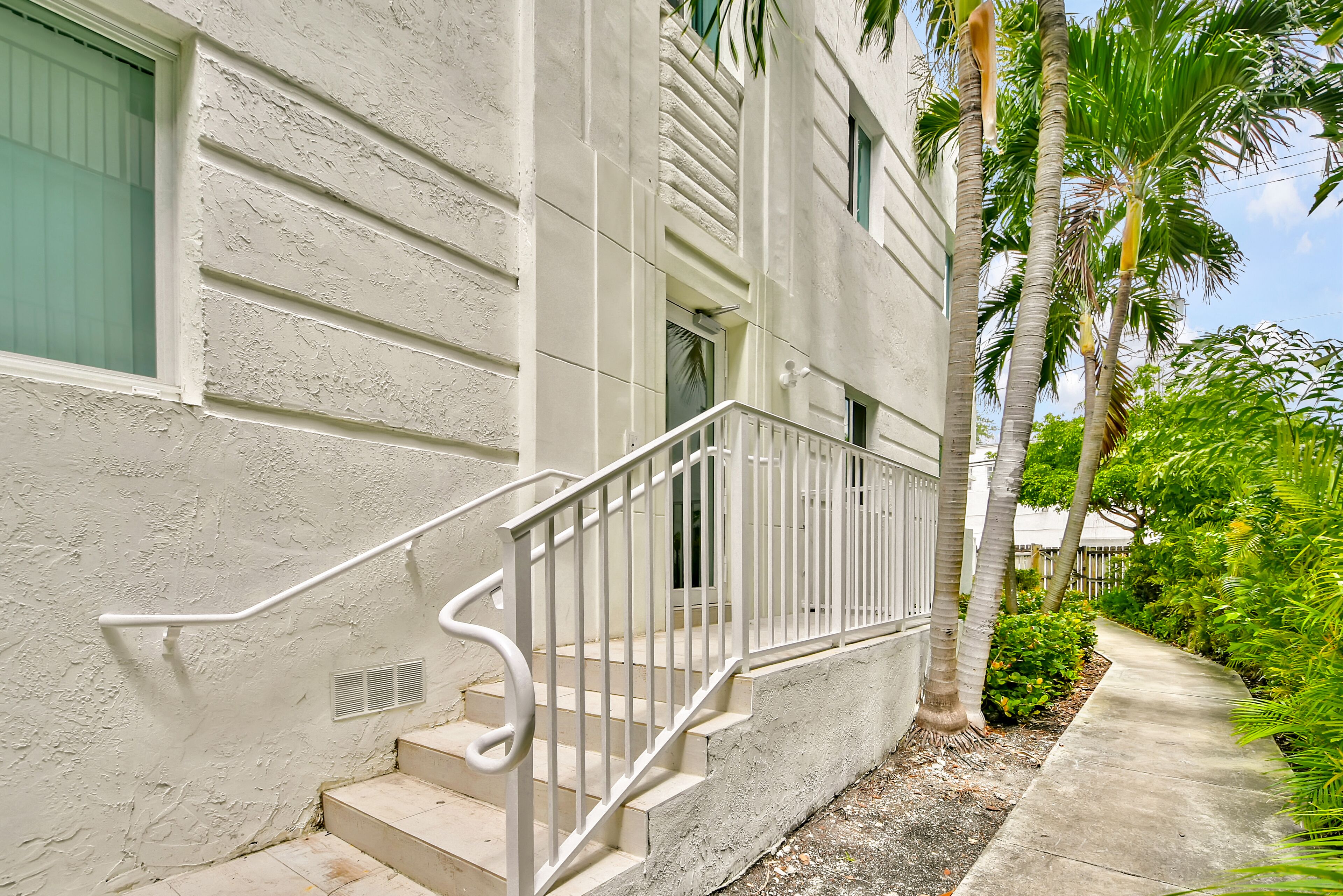 A building with a concrete exterior, a white railing on the steps, and a pathway lined with palm trees.