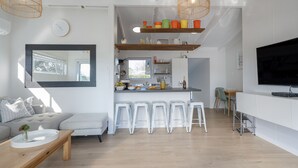 A modern living room with a sofa, coffee table, and a TV. There is a kitchen area with stools and a shelf with colorful items.