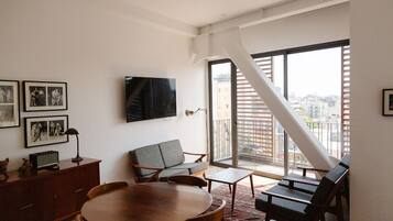 A spacious living room with a large TV, a dining table, and a balcony with a city view.