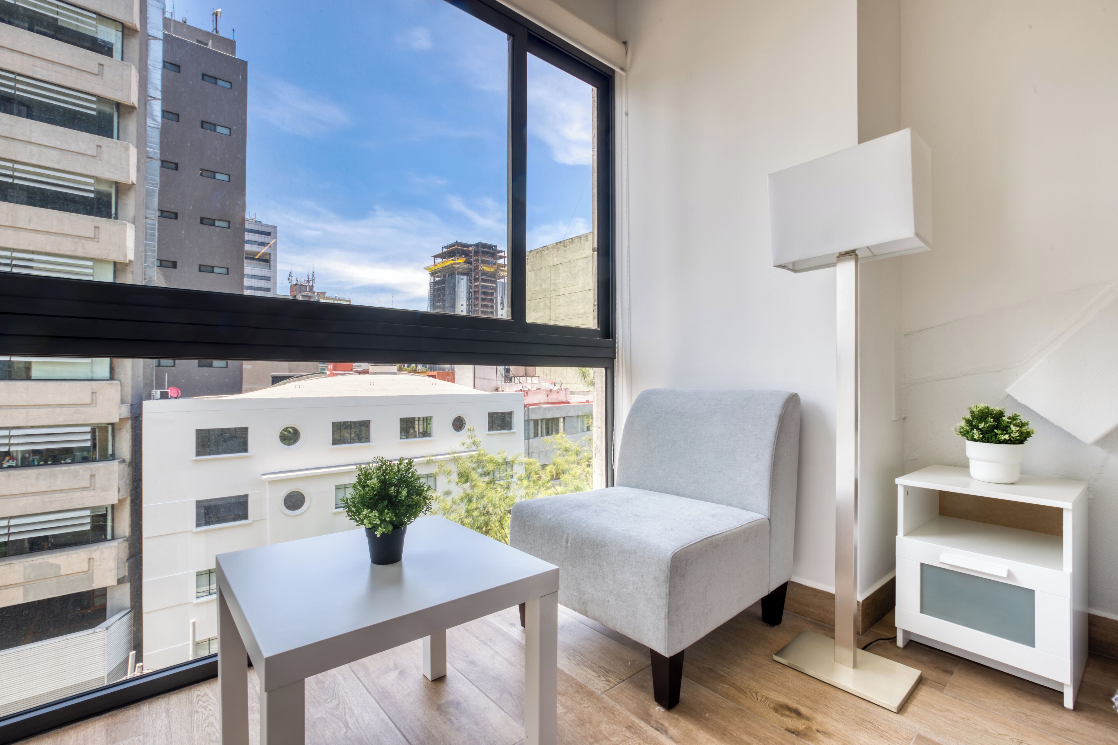 A modern apartment with a large window, a grey armchair, a small table with a plant, a white bedside table, and a floor lamp.