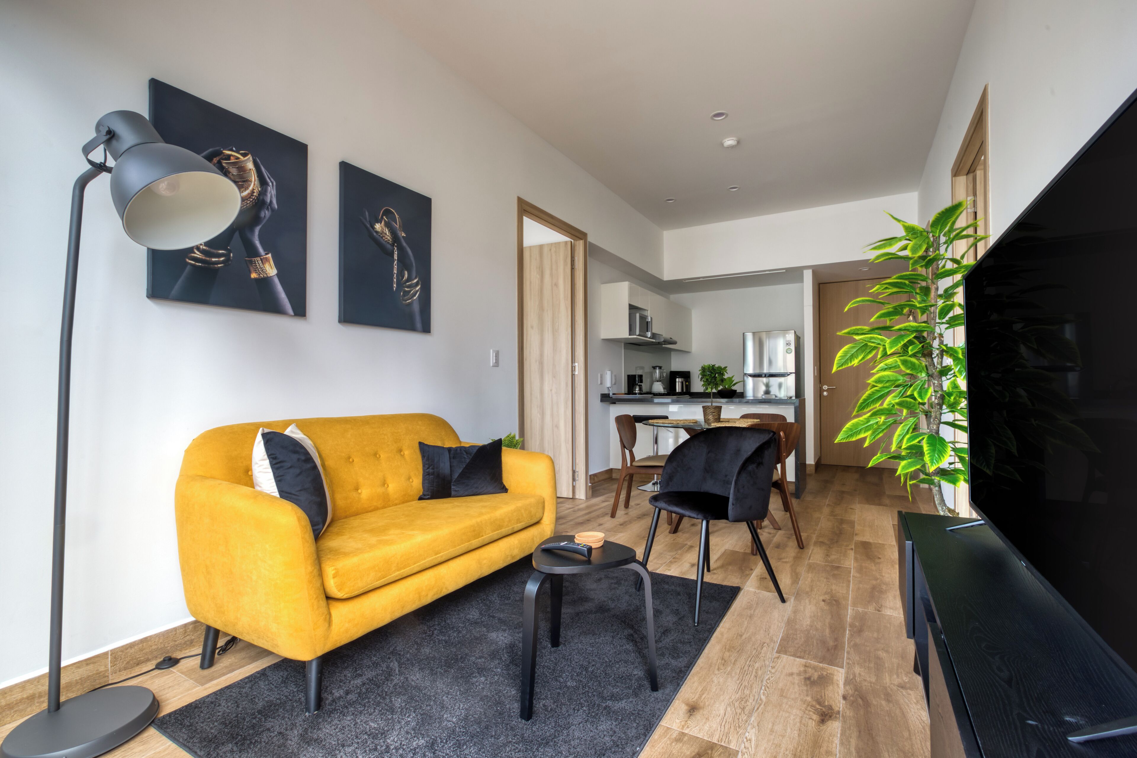 En moderne stue med en gul sofa, sorte lænestole, en gulvlampe i sort og et stort fladskærms-tv. Der er et spiseområde med bord og stole, og et køkken i baggrunden.