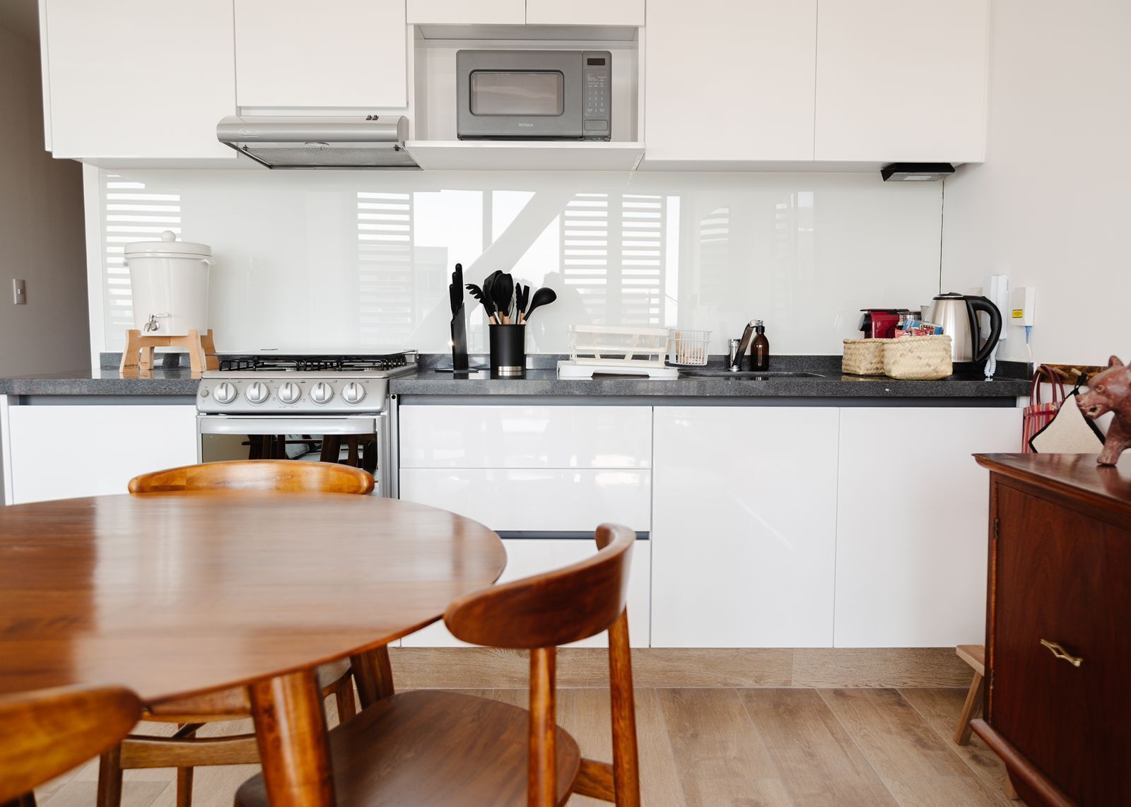 Una cocina moderna con armarios blancos, estufa a gas, microondas y comedor con mesa y sillas de madera.