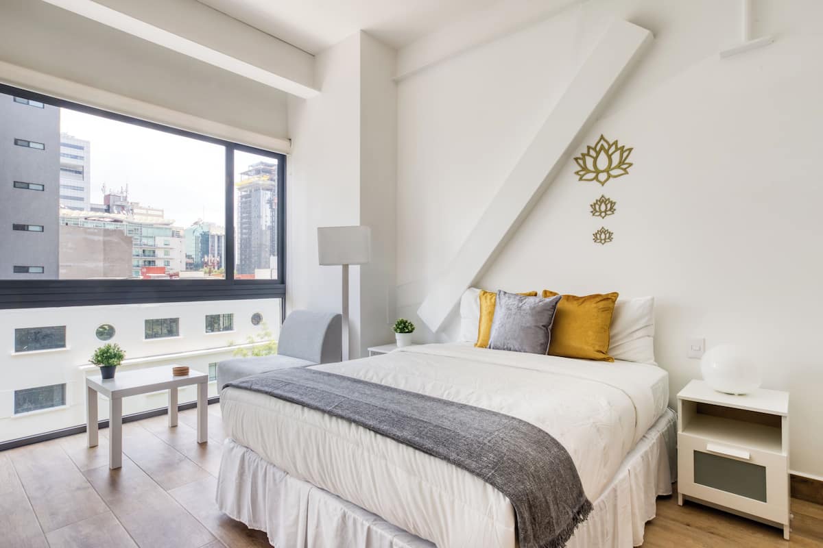 A modern bedroom with a large bed, a small bedside table, a nightstand, a window with a city view, and a decorative wall piece.