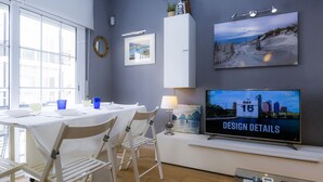 A modern dining area with a white table and chairs, a television displaying 'Day 16 Design Details', and a large window providing natural light. - SOLDUBA Apartment - A step away from the beach (Salou)