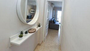 A hallway with a white shelf holding small plants, a large mirror, and a door leading to a living area with a gray sofa. - SOLIMAR Apartment - Only Families (Cambrils)