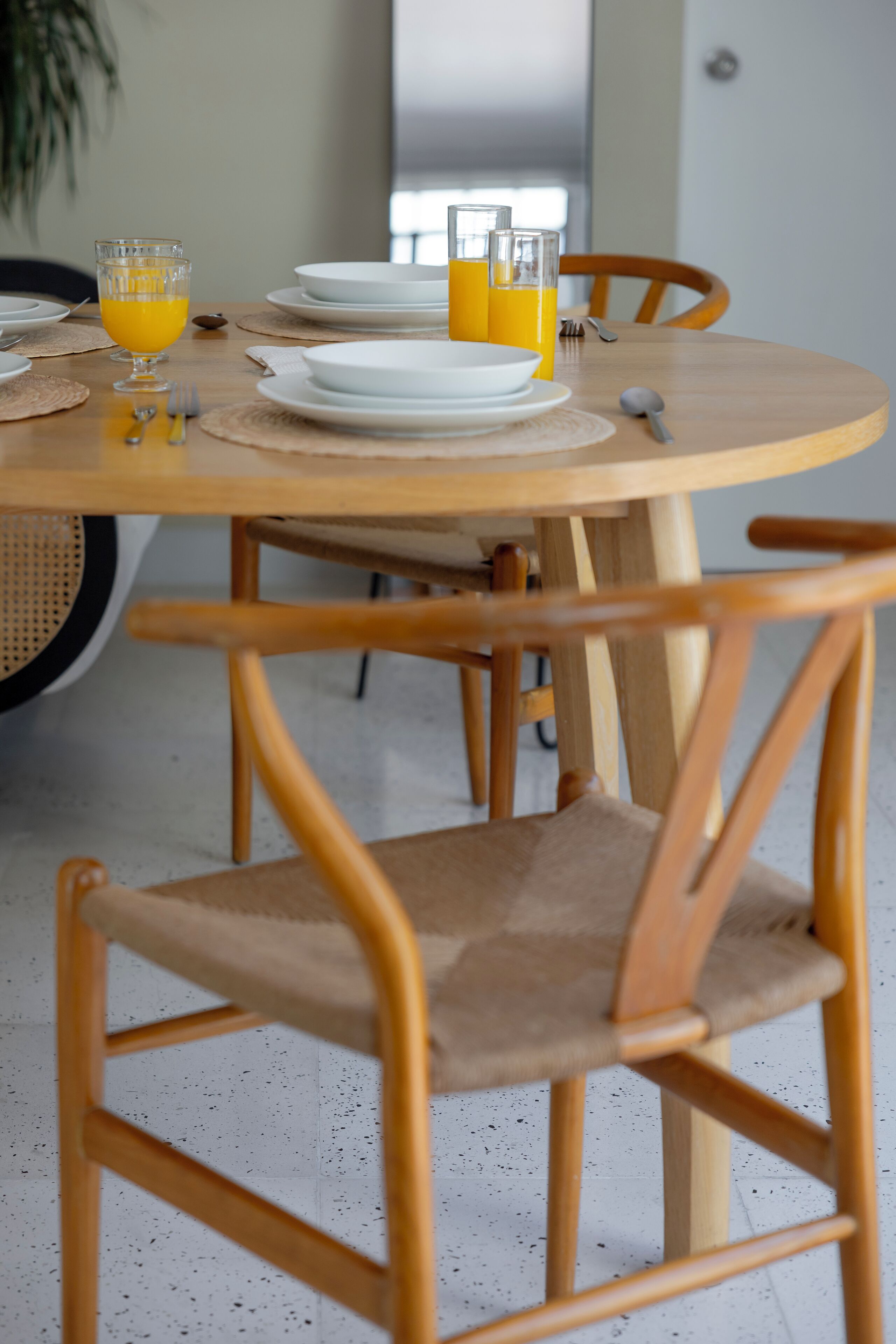 A round wooden dining table with white plates, glasses, and a bowl. There are two wooden chairs with cushioned seats.