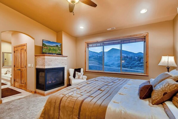Ein Schlafzimmer mit Kamin, großem Fenster und Bergblick, einem Bett mit gemusterten Betttextilien und einem Deckenventilator.