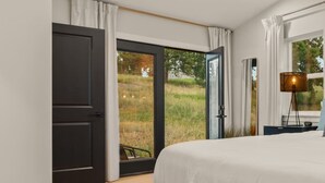 A bedroom with a large bed, a sliding glass door leading to a grassy outdoor area, and a lamp on a bedside table.