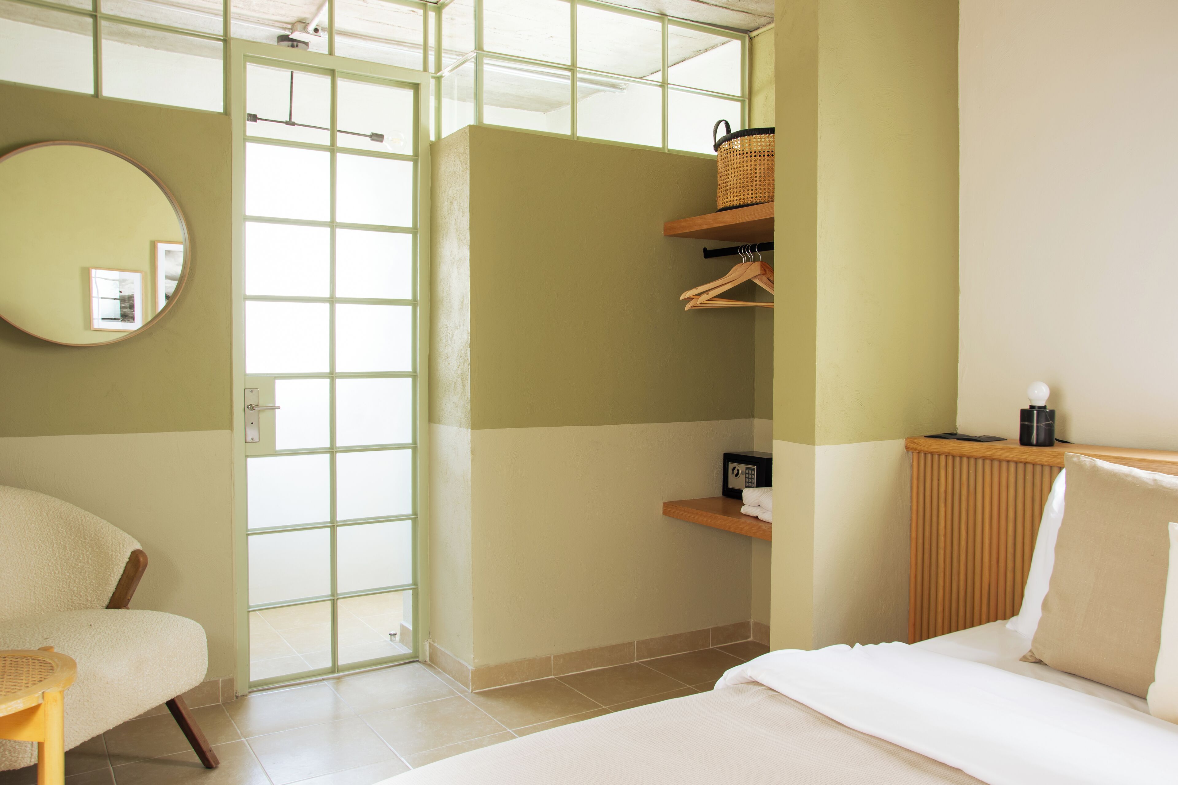 A hotel room with a bed, a chair, a round mirror, a wooden bedside table, a shelf with a basket, and a radiator.