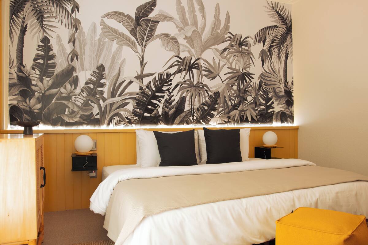 Habitación de hotel con una cama, dos mesitas de noche, un armario de madera y una pared cubierta de plantas tropicales.