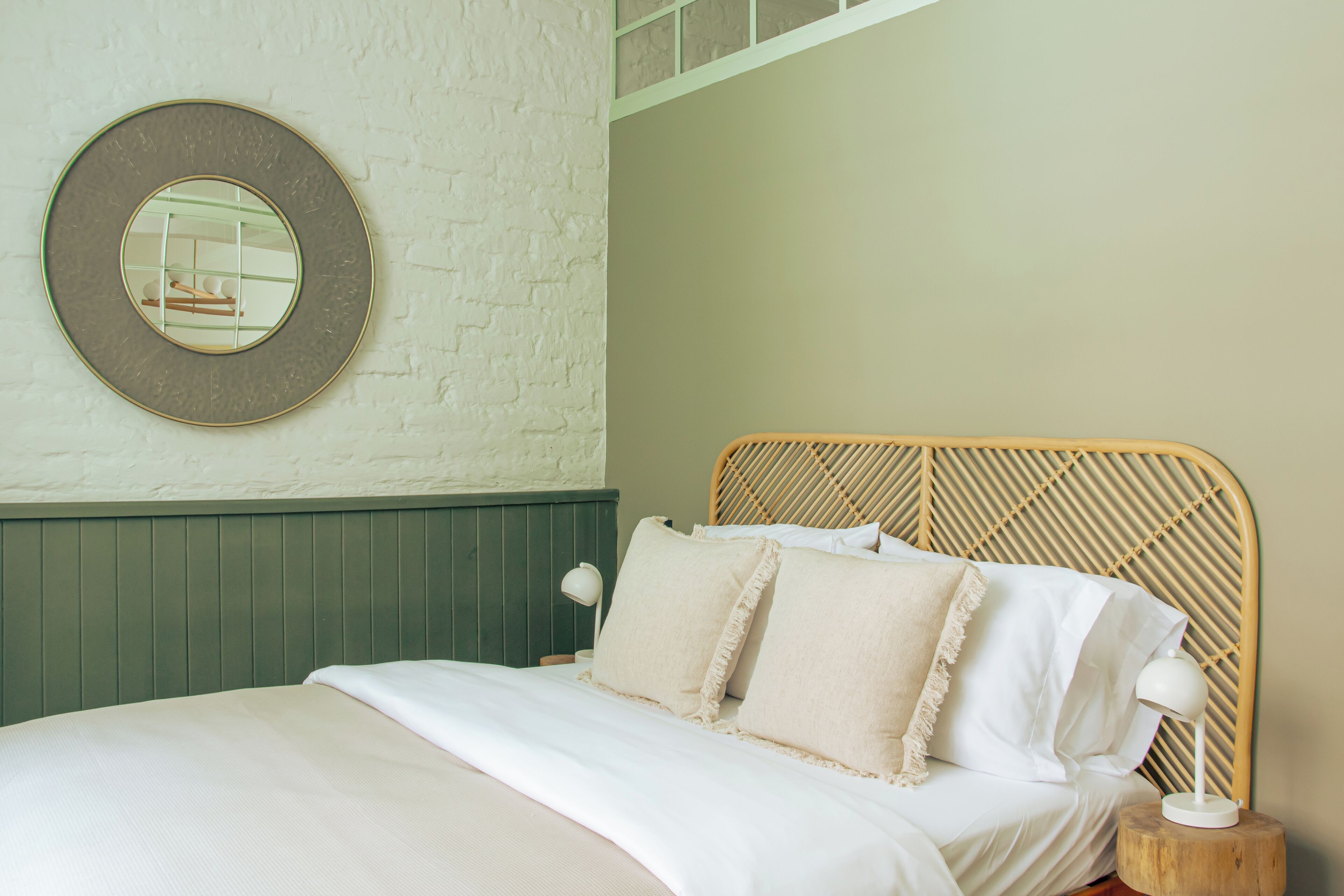 A neatly made bed with a wooden headboard, a bedside lamp, and a round mirror on the wall.