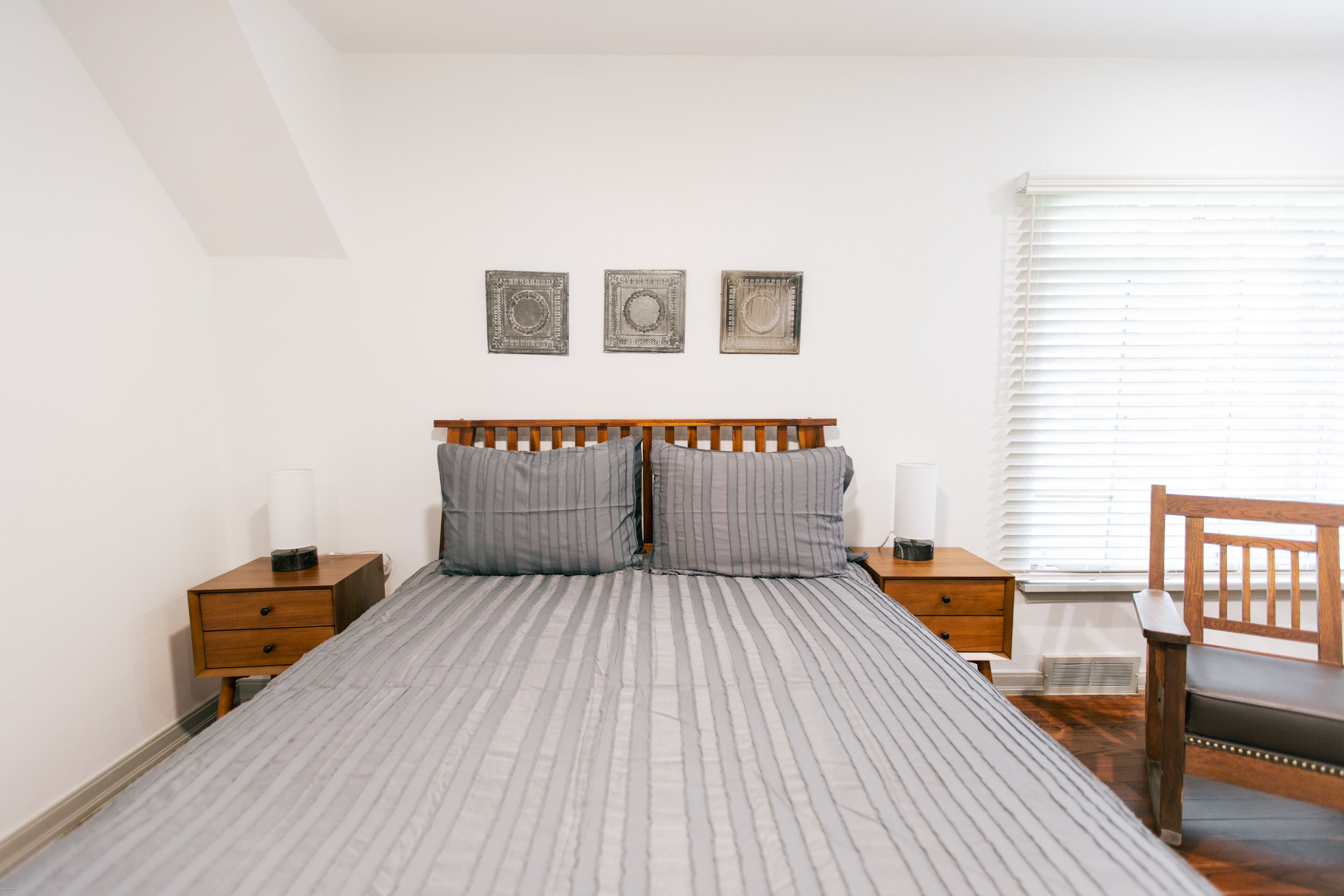 Une chambre bien rangée, avec un lit, deux tables de chevet, une chaise et une fenêtre avec des stores.