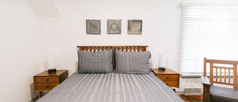 Une chambre bien rangée, avec un lit, deux tables de chevet, une chaise et une fenêtre avec des stores.