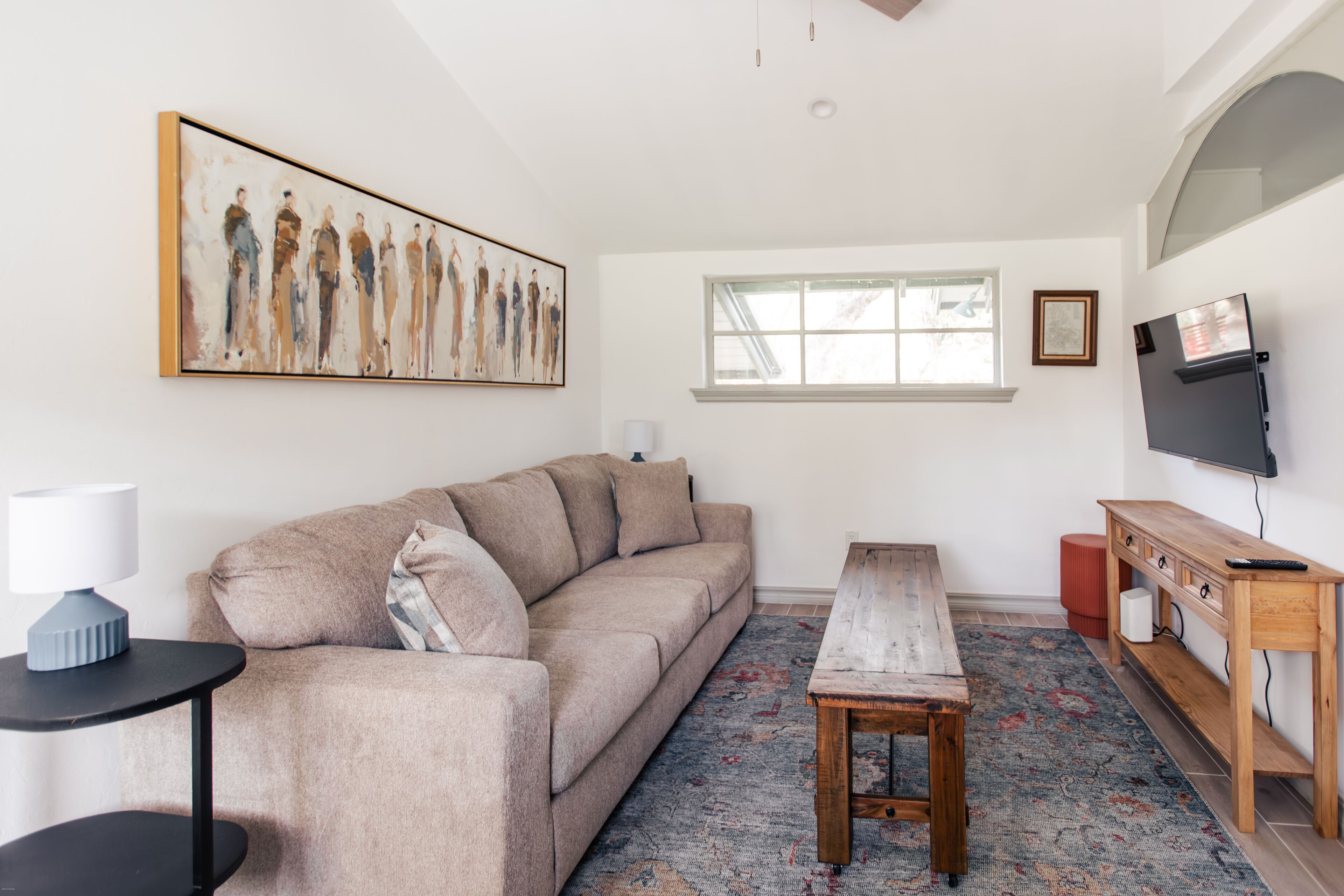 Una sala de estar con un sofá color beige, una mesa de centro de madera, un tapete estampado, un televisor en la pared y un cuadro sobre el sofá.