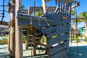A wooden play structure with climbing nets and a slide, surrounded by palm trees and a clear sky.