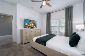 A bedroom with a bed, a TV on a dresser, a ceiling fan, a window with curtains, and a door.