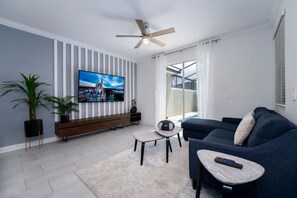 A modern living room with a flat-screen TV, a ceiling fan, a sofa, a coffee table, and a potted plant.