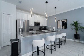 A modern kitchen with white cabinets, stainless steel appliances, a central island with bar stools, pendant lighting, and a potted plant.