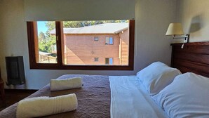A bedroom with a large window, a bed with white linens, a wooden headboard, and a wall-mounted lamp.