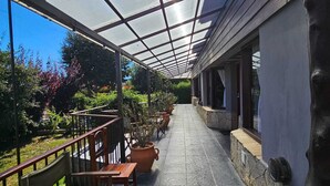 A covered outdoor area with a stone floor, wooden benches, potted plants, and a view of greenery.