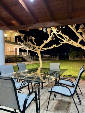 An outdoor patio with a glass table and chairs, a decorative tree, and a building in the background. - Villa Eva in Eretria Greece , the ideal place for your vacations (Eretria)