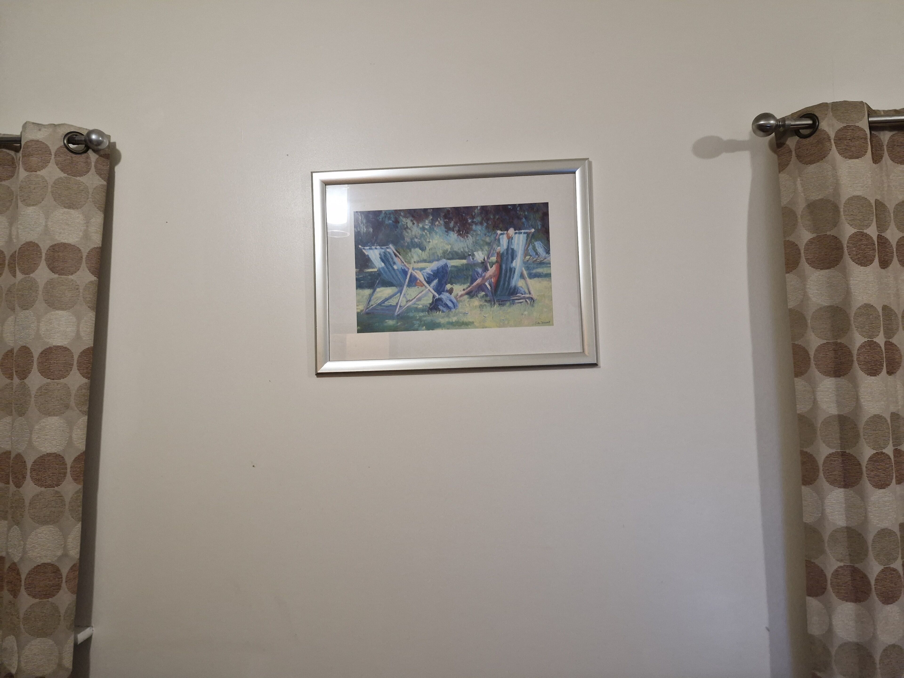 A framed painting of two people in a blue and white outdoor setting, hung on a plain wall between patterned curtains.